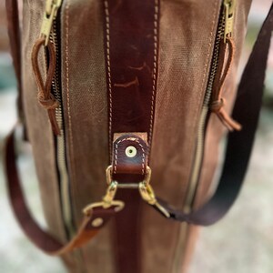 May include: A brown leather and canvas bag with a zipper closure and a brown leather strap. The bag has a gold metal buckle and a gold metal zipper pull.