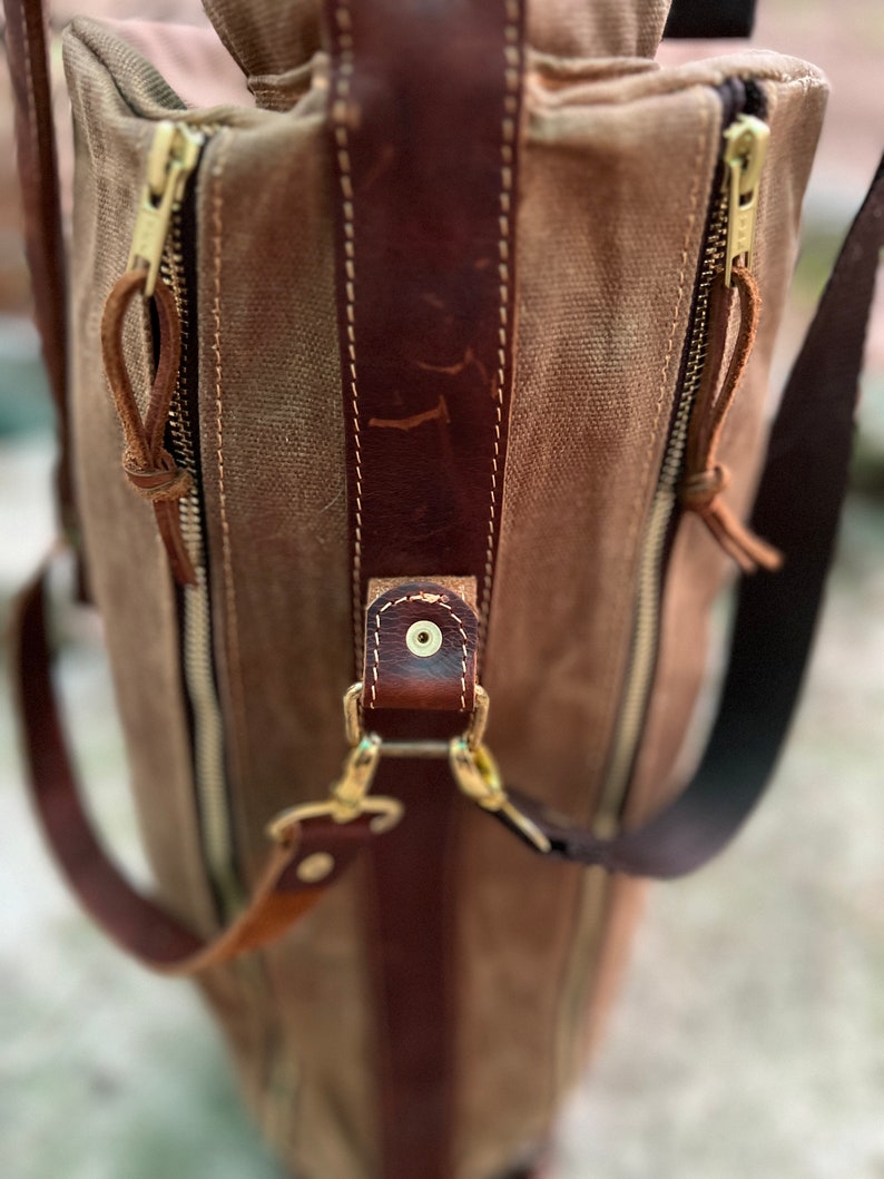 May include: A brown leather and canvas bag with a zipper closure and a brown leather strap. The bag has a gold metal buckle and a gold metal zipper pull.
