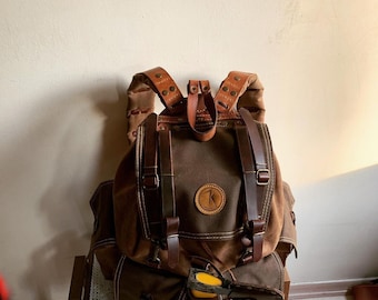 May include: A brown canvas backpack with leather straps and buckles. The backpack has a large main compartment and a smaller front pocket. The backpack is decorated with a leather patch with a letter "K" on it.