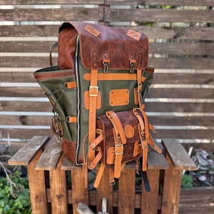 Puede incluir: Una mochila con múltiples compartimentos hecha de cuero verde oliva y cuero marrón desgastado. Cuenta con una solapa superior grande, un bolsillo frontal más pequeño, correas de cuero beige ajustables con hebillas de latón y bolsillos laterales.