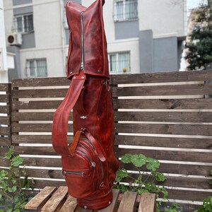 Puede incluir: Una bolsa de golf de cuero marr&oacute;n rojizo con m&uacute;ltiples compartimentos con cremallera y una correa para el hombro. La bolsa tiene un perfil alto y delgado y est&aacute; hecha de cuero texturizado. La bolsa est&aacute; sobre un pal&eacute; de madera frente a una valla de madera.