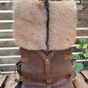 Può includere: Uno zaino in pelle marrone con patta in pelliccia. Lo zaino ha spallacci regolabili in pelle e tasche laterali. La patta in pelliccia è marrone chiaro con una striscia marrone scuro al centro.