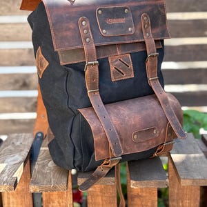 Puede incluir: Mochila de lona negra con detalles en cuero marrón. La mochila tiene cierre de solapa, correas ajustables y un bolsillo frontal. El cuero tiene un aspecto envejecido y la bolsa parece resistente. La mochila está sobre una superficie de madera.