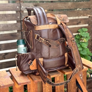 May include: A brown leather backpack with a large front pocket and a smaller pocket on the side. The backpack has a green insulated water bottle in the side pocket. The backpack is sitting on a wooden surface.