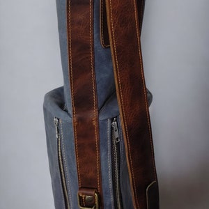 May include: A gray canvas bag with brown leather straps and zippers. The bag has a top handle and a shoulder strap.