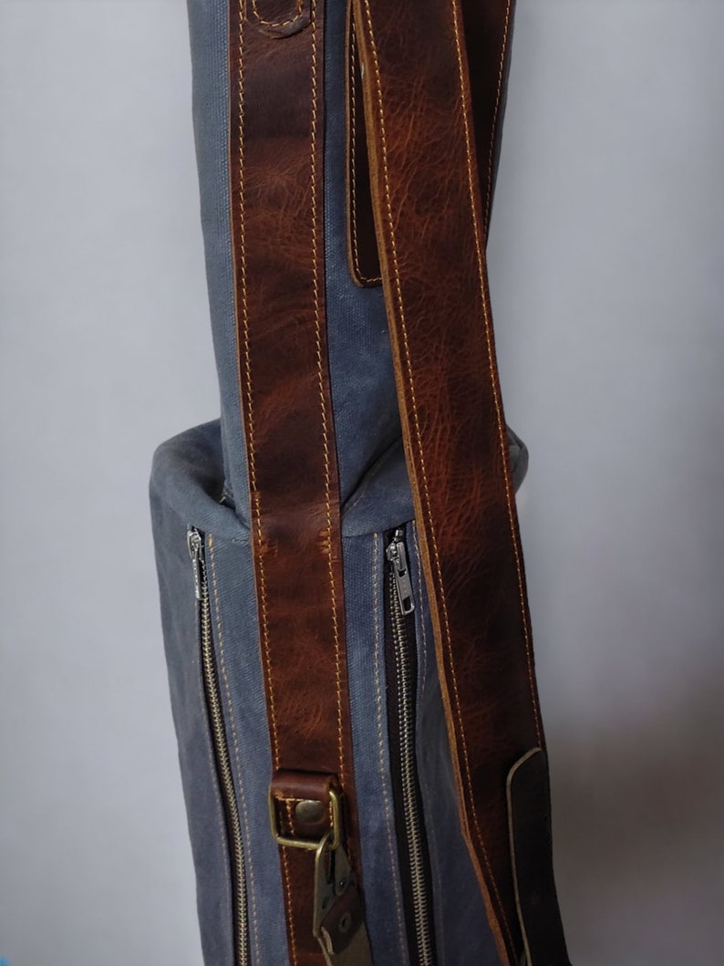 May include: A gray canvas bag with brown leather straps and zippers. The bag has a top handle and a shoulder strap.