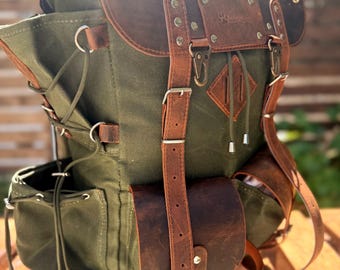 Könnte beinhalten: Ein grüner Canvas-Rucksack mit braunen Lederakzenten. Der Rucksack verfügt über mehrere Taschen, verstellbare Riemen und eine obere Klappe mit Reißverschluss. Das Leder hat einen Used-Look, und die Hardware ist silberfarben. Die Tasche ist für den Außenbereich konzipiert.