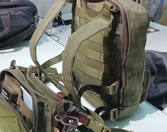May include: A green canvas backpack with multiple pockets and straps. The backpack has a brown leather handle and zipper closures. A smaller green canvas bag with a brown leather strap and multiple pockets is attached to the backpack.