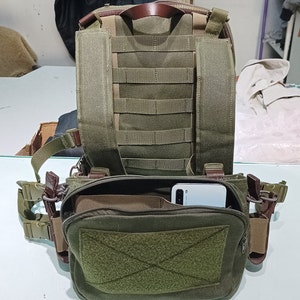 May include: A green canvas rucksack with a large zipped pocket on the front. The rucksack has a brown leather handle and adjustable straps. A white smartphone is visible inside the front pocket.