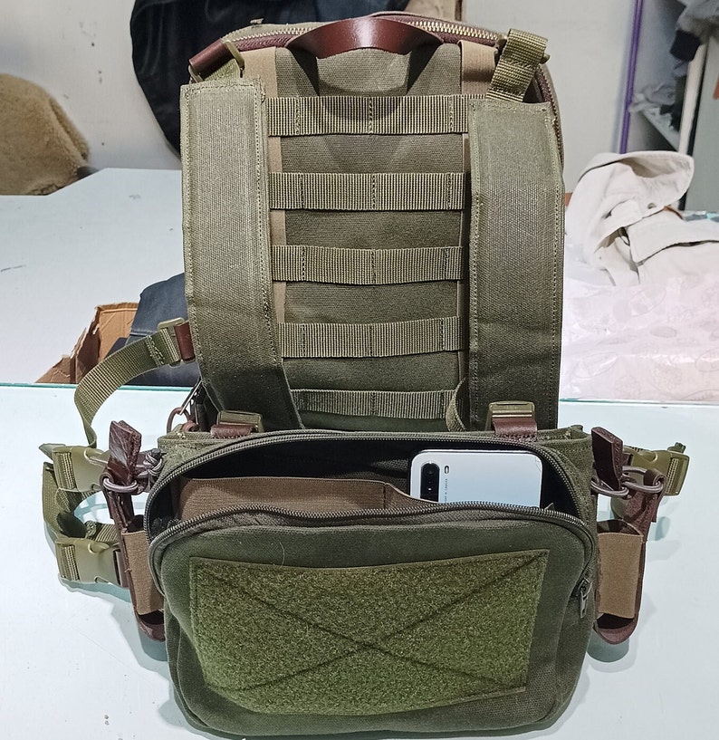 May include: A green canvas rucksack with a large zipped pocket on the front. The rucksack has a brown leather handle and adjustable straps. A white smartphone is visible inside the front pocket.