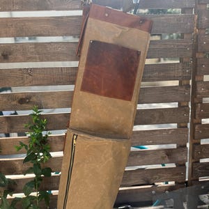 May include: A tan golf club bag with a brown leather top and pocket, hanging on a wooden fence. The bag has a zippered pocket and holds two golf clubs. The bag is made of canvas material.