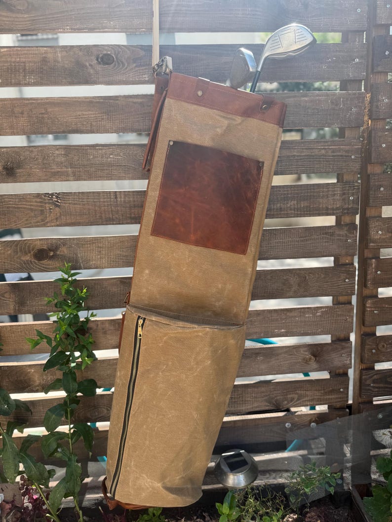 May include: A tan golf club bag with a brown leather top and pocket, hanging on a wooden fence. The bag has a zippered pocket and holds two golf clubs. The bag is made of canvas material.