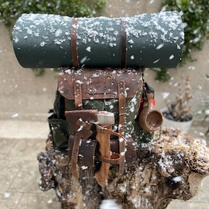 May include: A green canvas backpack with leather straps and buckles. The backpack is filled with camping gear, including a sleeping mat, a small axe, and a wooden cup. The backpack is sitting on a wooden stump covered in snow.