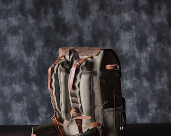May include: A brown leather and canvas backpack with adjustable straps and buckles. The backpack has a large main compartment and several smaller pockets.