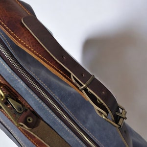 May include: A blue canvas bag with a brown leather strap and buckle. The bag has a zipper closure and a brown leather trim.