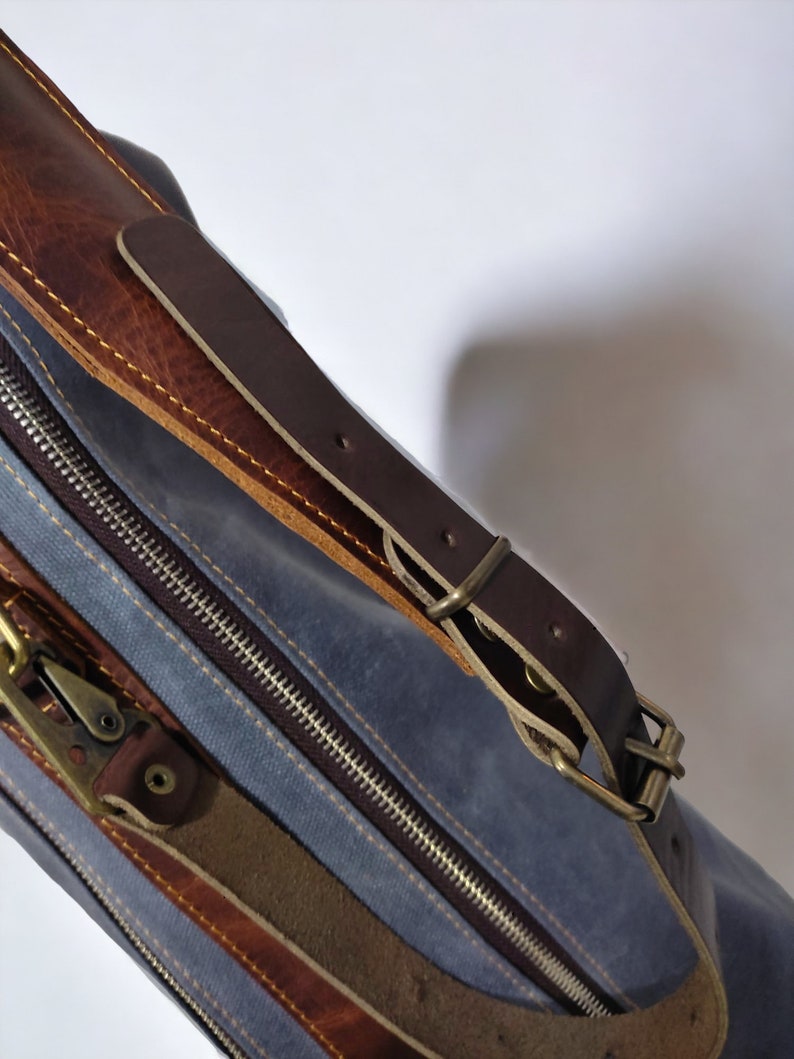 May include: A blue canvas bag with a brown leather strap and buckle. The bag has a zipper closure and a brown leather trim.