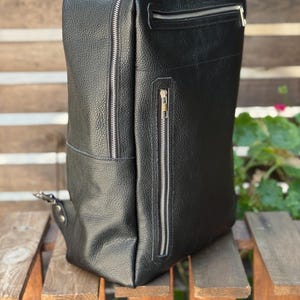 May include: A black textured leather backpack featuring a vertical zippered pocket on the front and a horizontal zippered pocket at the top. The bag has silver-toned metal zippers and a structured rectangular shape with a side pocket.