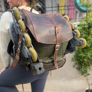 Könnte beinhalten: Ein brauner Leder- und grüner Canvas-Rucksack mit einer Reißverschlusstasche und einem Riemen. Der Rucksack hat ein Paar Inline-Skates daran befestigt.