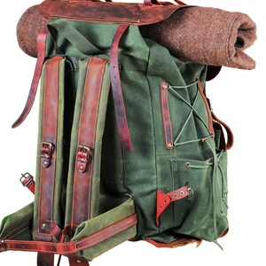 May include: A green canvas backpack with brown leather straps and buckles. The backpack has a roll-top closure and a flap with a buckle. The backpack is empty and has a brown wool blanket on top.