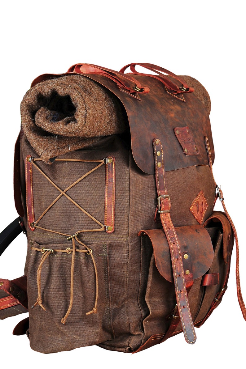 May include: A brown leather and canvas backpack with a rolled up blanket inside. The backpack has a large front pocket with a leather flap and buckle closure. The backpack has a brown leather strap with a buckle closure.