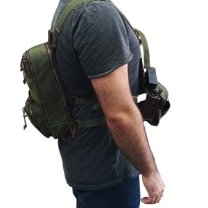 May include: A green canvas backpack with multiple pockets and straps. The backpack is being worn by a person wearing a dark shirt and blue jeans.