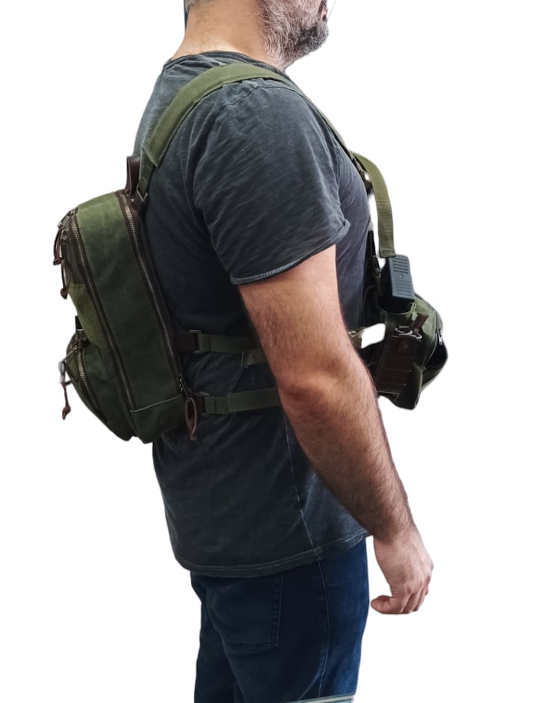 May include: A green canvas backpack with multiple pockets and straps. The backpack is being worn by a person wearing a dark shirt and blue jeans.