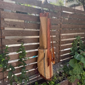 May include: A tan golf club bag with a brown leather shoulder strap and accents hangs on a wooden fence. The bag has a full-length zip and holds a golf club. The background includes greenery and a building.