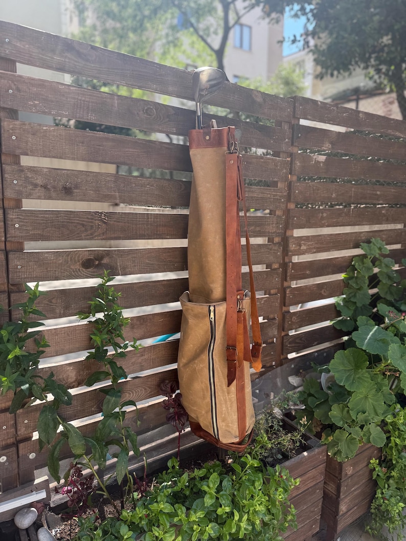 May include: A tan golf club bag with a brown leather shoulder strap and accents hangs on a wooden fence. The bag has a full-length zip and holds a golf club. The background includes greenery and a building.