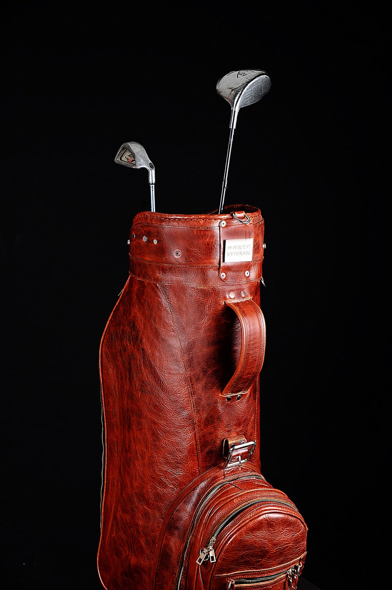 Może przedstawiać: Worek golfowy w stylu vintage, wykonany ze sk&oacute;ry w kolorze czerwonobrązowym, z dwoma kijami golfowymi. Torba posiada uchwyt, kieszeń zapinaną na zamek oraz metalową tabliczkę z tekstem. Kije golfowe są srebrne i czarne. Torba stoi na czarnym tle.