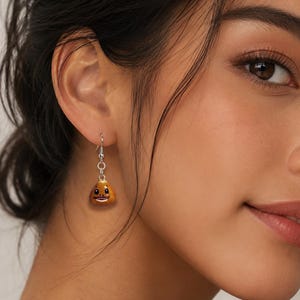 May include: A drop earring featuring a small, brown, glossy, rounded charm with a hand-painted face. The charm is suspended from a silver-toned metal hook and a short chain link, creating a dangling design that hangs from the earlobe.