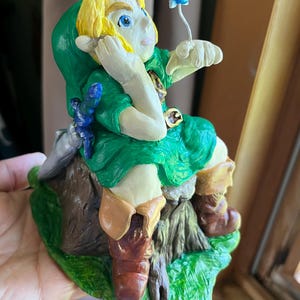 May include: A hand-painted figurine of a blonde character in a green tunic and hood, sitting on a brown tree stump. The character has a blue sword at their side and is looking at a small, light blue fairy hovering on a thin wire above their hand.