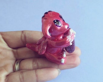 Op de afbeelding: Een klein, roze, glinsterend drakenbeeldje met witte accenten en paarse ogen. De draak houdt een hart in zijn poten.