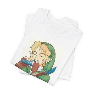 May include: A white t-shirt featuring a watercolour-style illustration of a blonde character with pointed ears wearing a green tunic and brown gloves, playing a blue ocarina. The character has a blue sword hilt visible over their shoulder.