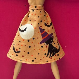 May include: A doll skirt in a light orange fabric featuring a pattern of black and orange polka dots, black bats, a white full moon, and a black cat wearing a purple and orange witch hat. The skirt has a fitted waistband and flared hem.