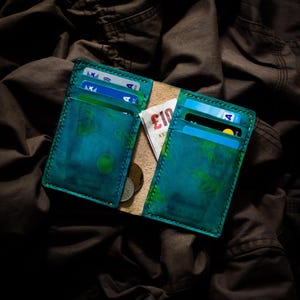 May include: Open leather wallet with a teal and green watercolour design. The wallet contains credit cards, cash, and coins. The wallet is on a dark brown fabric background.