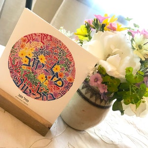 May include: A colorful card with a circular design featuring Hebrew text and floral elements. The card is displayed on a wooden stand and is next to a vase of flowers.
