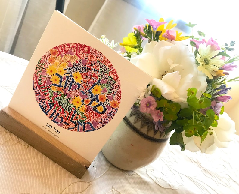 May include: A colorful card with a circular design featuring Hebrew text and floral elements. The card is displayed on a wooden stand and is next to a vase of flowers.