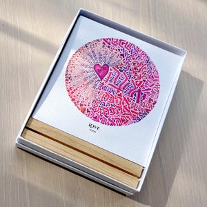 May include: A white card with a pink and purple circle design featuring Hebrew text and a pink heart in the center. The card is in a white box with a wooden base. The text on the card reads "LOVE" and "אהבה".