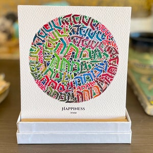 May include: A white card with a colorful circular design made of Hebrew letters. The card is in a white box and the text "HAPPINESS" and "שמחה" are printed below the design.