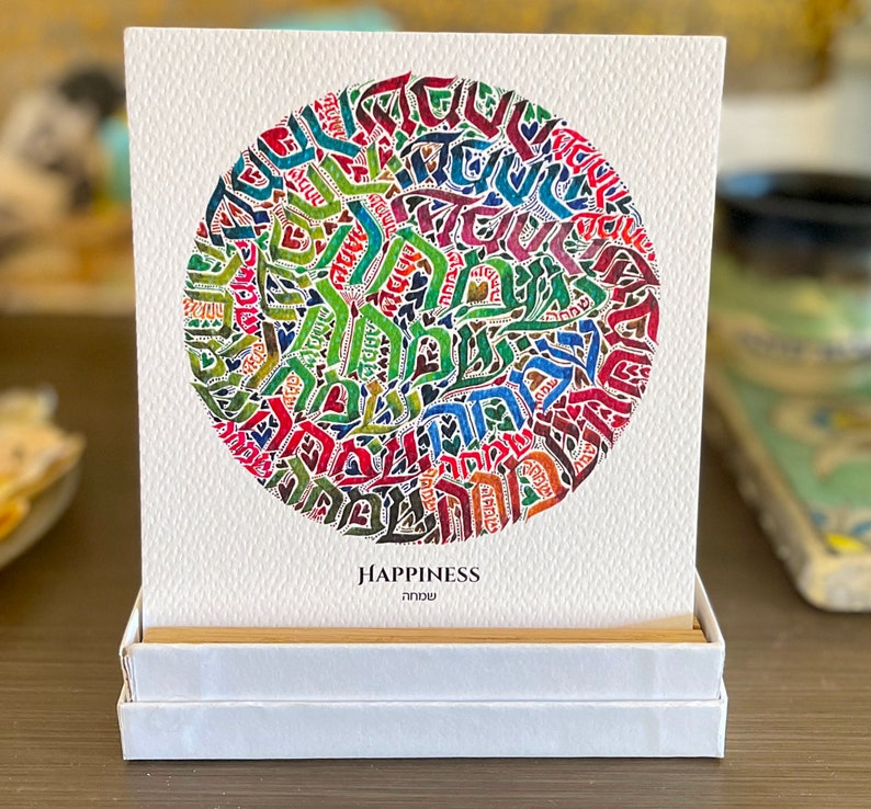 May include: A white card with a colorful circular design made of Hebrew letters. The card is in a white box and the text "HAPPINESS" and "שמחה" are printed below the design.