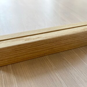 May include: A natural wood display stand with a single slot for holding cards or small items. The stand is made of light brown wood with a smooth finish.