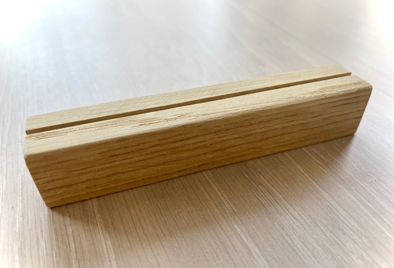 May include: A natural wood display stand with a single slot for holding cards or small items. The stand is made of light brown wood with a smooth finish.