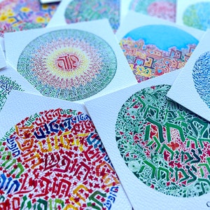 May include: A collection of colorful circular art prints featuring Hebrew calligraphy. The prints are on white cardstock and have a variety of designs, including a mandala, a tree, and a geometric pattern. The text on one of the prints reads "GROWTH".
