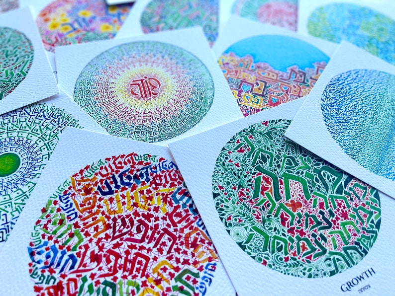 May include: A collection of colorful circular art prints featuring Hebrew calligraphy. The prints are on white cardstock and have a variety of designs, including a mandala, a tree, and a geometric pattern. The text on one of the prints reads "GROWTH".