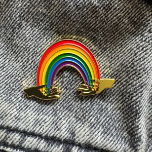 Peut inclure: Une épingle en émail arc-en-ciel colorée avec un contour doré. L'arc-en-ciel est au-dessus de deux mains dorées, avec des étoiles et des couleurs qui s'écoulent des extrémités. L'épingle est posée sur un fond gris texturé.