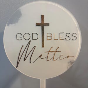 May include: A white, round cake topper with the words "GOD BLESS Matter" and a cross design. The text and cross are in a gold color. The topper has a small handle at the bottom.