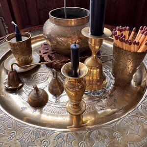 May include: A large round brass tray featuring intricate etched floral patterns, holding a collection of brass items including two candle holders with black candles, a small brass bowl, a brass cup filled with wooden matches, and two small brass bells.