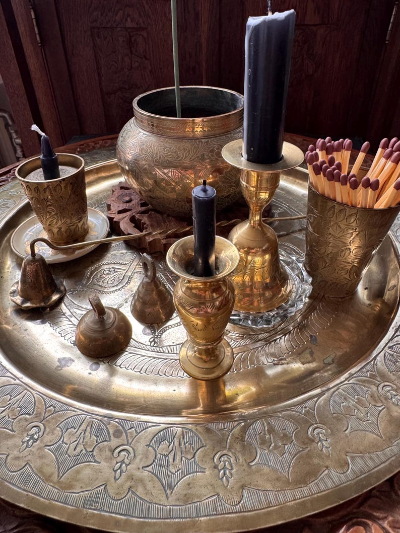 May include: A large round brass tray featuring intricate etched floral patterns, holding a collection of brass items including two candle holders with black candles, a small brass bowl, a brass cup filled with wooden matches, and two small brass bells.