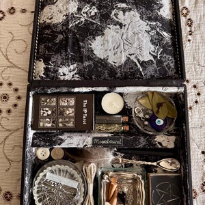 Op de afbeelding: Open zwarte doos met een zwart-wit patroon aan de binnenkant, met tarotkaarten, kaarsen, kleine flesjes, een lepel en andere mystieke items. De doos is gevoerd met een zwart-witte stof met een gedetailleerd ontwerp.