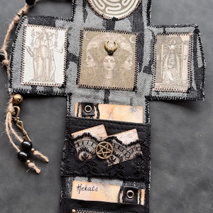 Può includere: Un porta carte dei tarocchi fatto a mano, a forma di croce, con un motivo in denim nero e grigio. Presenta vari simboli occulti, tra cui un labirinto, una luna crescente e un pentagramma. La parola "Hekate" è scritta sopra.